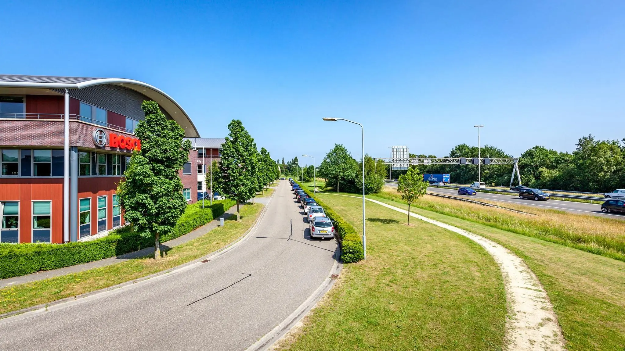 Kantoor van Bosch aan een rustige weg met geparkeerde auto's, naast een snelweg en een grasveld met wandelpad.